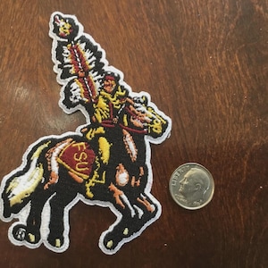 Può includere: Toppa ricamata raffigurante un guerriero Seminole a cavallo, con le lettere "FSU" sulla sella. La toppa è nera, oro e rossa, con bordi bianchi. Una moneta è accanto per il confronto delle dimensioni.