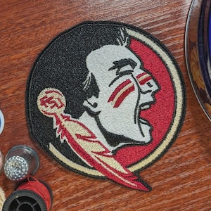 Può includere: Toppa ricamata con un profilo di capo nativo americano in nero, bianco e rosso. La toppa circolare ha un bordo rosso e un'emblema di piuma. Le lettere "FSU" sono visibili sulla piuma.