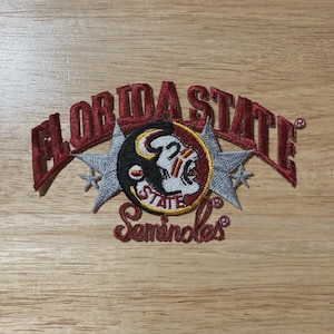 Può includere: Toppa ricamata bordeaux con le parole "FLORIDA STATE" arcuate sopra un emblema circolare. L'emblema include una testa di Seminole e la parola "STATE". Due stelle argentate affiancano l'emblema, con "Seminoles" sotto.
