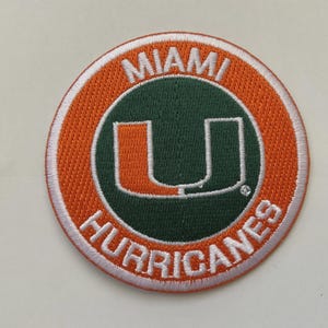 May include: Circular embroidered patch with the Miami Hurricanes logo. The patch has an orange border and white stitching. The centre features a green background with a white "U" and the words "MIAMI HURRICANES" in white.