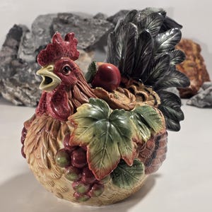 May include: A ceramic rooster-shaped container with a red comb and wattle, a yellow beak, and a black and gray tail. The body is adorned with green leaves and red grapes. The container is approximately 8 inches in height.