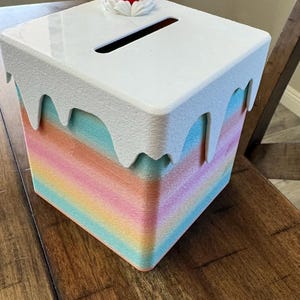 May include: A square, pastel-colored box with a white, dripping-icing top. A red strawberry with green leaves sits atop the box. The box has a slot for inserting items, and is likely a decorative storage container.