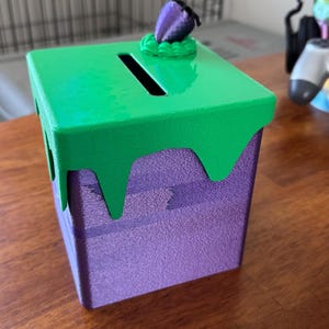 Può includere: Una scatola decorativa viola e verde con una fessura sulla parte superiore. La parte superiore verde ha un effetto di vernice gocciolante. Una piccola bacca viola è in cima. La scatola è su una superficie di legno.