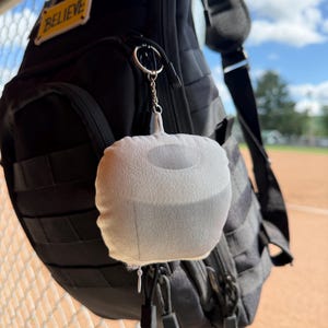 May include: A black backpack with multiple compartments and straps hangs on a metal fence. A small, gray plush keychain in the shape of a sphere is attached to the backpack. The word "BELIEVE" is on a patch on the backpack.