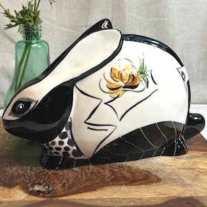 Könnte beinhalten: Keramik-Kaninchenfigur mit schwarz-weißem Design und floralem Akzent. Das Kaninchen steht auf einem Holzschneidebrett. Eine kleine grüne Glasvase und rosa Blumen befinden sich im Hintergrund.