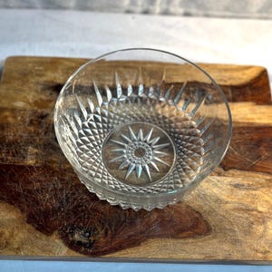 May include: A clear glass bowl featuring a starburst design and diamond patterns. The bowl sits on a wooden chopping board with a natural wood grain pattern. The bowl is likely for serving.
