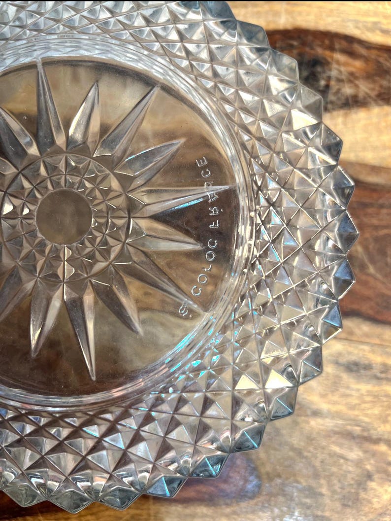 May include: Clear glass dish with a starburst design in the centre and a geometric pattern around the rim. The dish has the text "S. COLOC FRANCE" etched on the side.