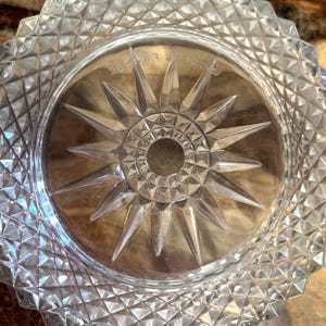 May include: Clear glass dish with a starburst design in the centre. The dish has a diamond-cut pattern around the rim. The starburst has a central hole and pointed rays. The dish is on a wooden surface.
