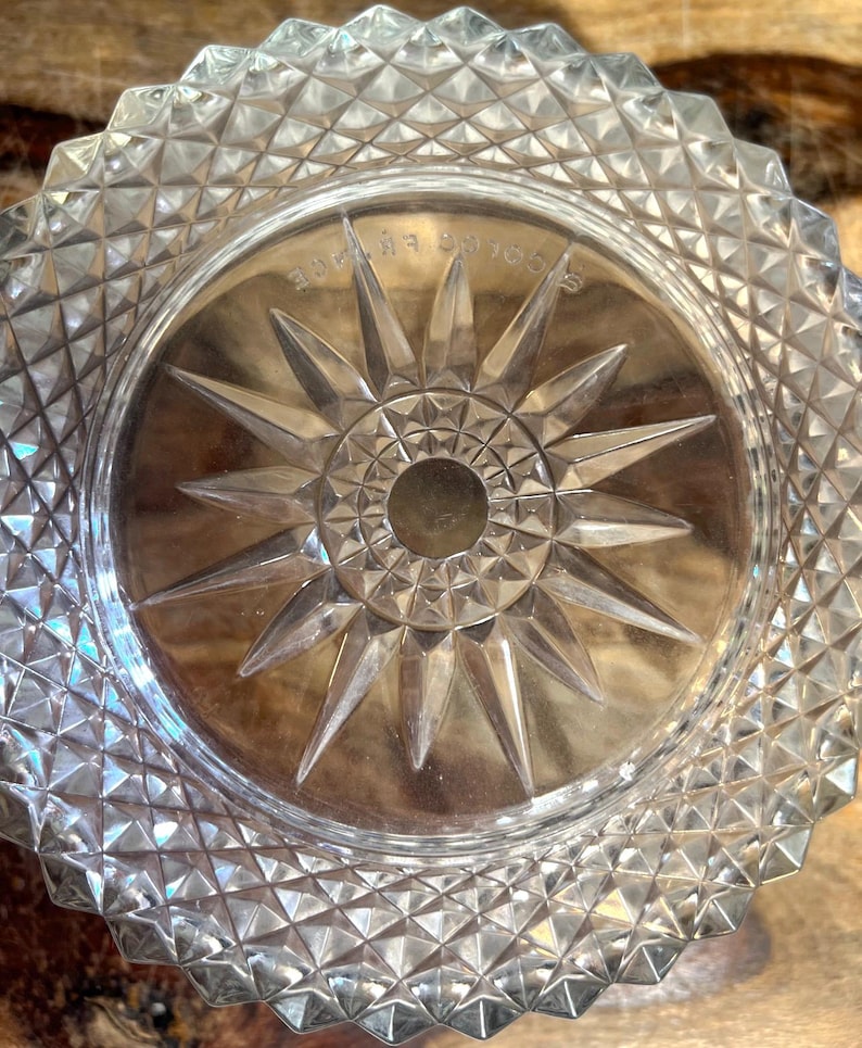 May include: Clear glass dish with a starburst design in the centre. The dish has a diamond-cut pattern around the rim. The starburst has a central hole and pointed rays. The dish is on a wooden surface.