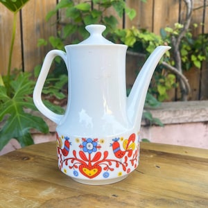 Puede incluir: Cafetera de cerámica blanca con un pico largo y asa. La cafetera presenta un colorido diseño de arte popular con pájaros, flores y corazones en rojo, azul, amarillo y naranja. La cafetera tiene tapa y está sobre una superficie de madera.