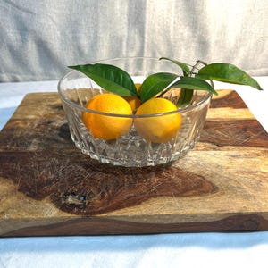 May include: A clear glass bowl with a decorative pattern contains three bright orange oranges and green leaves. The bowl rests on a rustic wooden chopping board. The background is light-coloured.