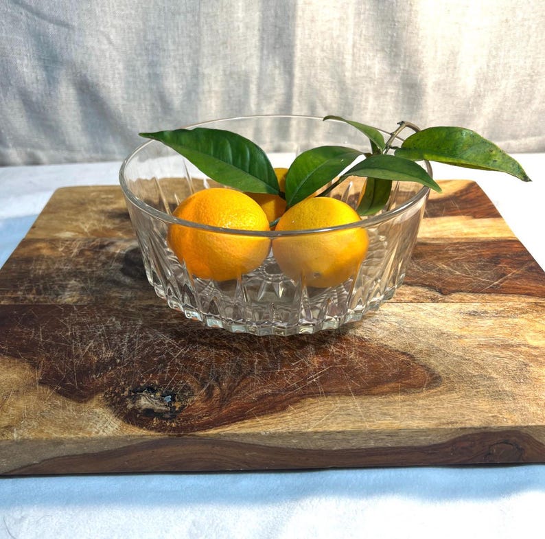 May include: A clear glass bowl with a decorative pattern contains three bright orange oranges and green leaves. The bowl rests on a rustic wooden chopping board. The background is light-coloured.