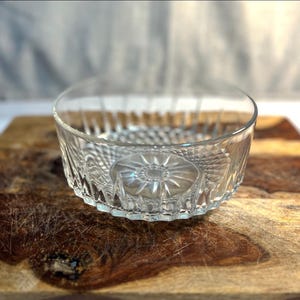 May include: A clear, round glass bowl with a decorative, textured pattern. The bowl features a starburst design on the base and a cut-glass rim. The bowl is on a wooden chopping board.
