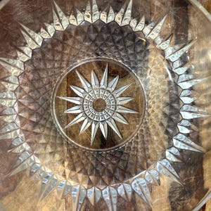 May include: Clear glass plate featuring a starburst design in the centre and around the rim. The plate has a textured surface and is set against a wooden background. The starburst pattern is repeated throughout.