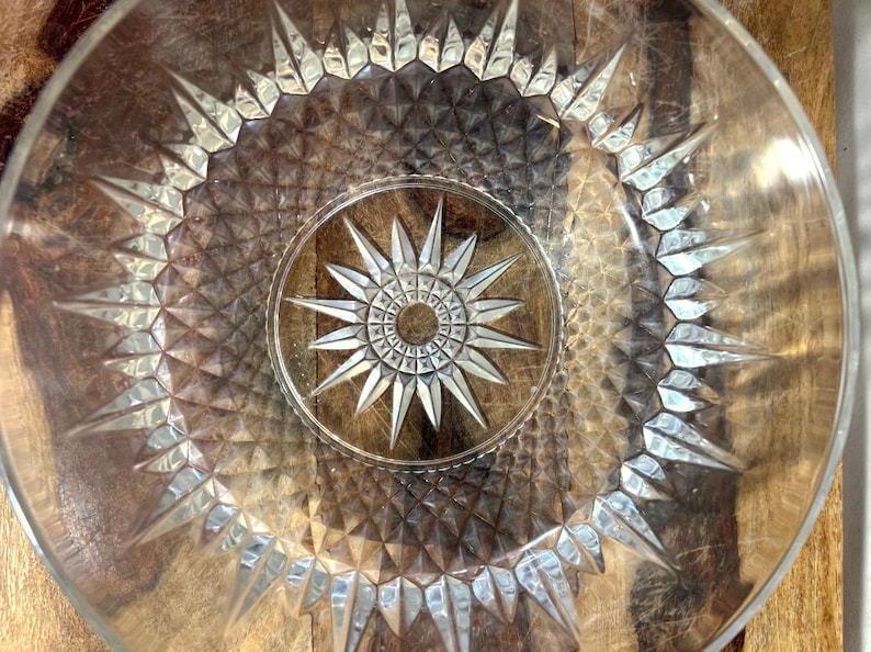 May include: Clear glass plate featuring a starburst design in the centre and around the rim. The plate has a textured surface and is set against a wooden background. The starburst pattern is repeated throughout.