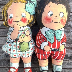 May include: Two vintage fabric dolls. A girl doll with blonde hair, a blue bow, and a floral dress. The boy doll wears a red and white striped outfit with a bow tie. Both dolls have rosy cheeks and painted facial features, reminiscent of a bygone era.