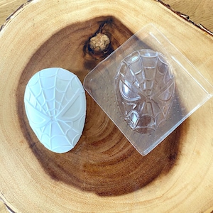 May include: A white, oval-shaped mold with a spiderweb design next to a clear plastic mold in the shape of a superhero mask. The items are on a wooden surface with a natural wood grain.