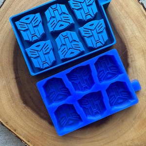 May include: Two blue silicone ice cube trays. Each tray has six moulds shaped like a robot face. The trays are on a wooden surface. The ice cube trays are a fun and unique way to make ice cubes.