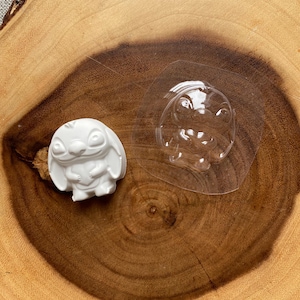 May include: A white, three-dimensional Stitch figurine next to a clear plastic mold of the same character. The figurine is on a wooden surface with a natural wood grain pattern. The figurine is a popular character from the animated film Lilo & Stitch.