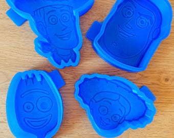 May include: Four bright blue plastic cookie cutters featuring the faces of four distinct animated characters. Each cutter has a raised outline and internal details to imprint the character's features into dough, including a cowboy, a space ranger, a spork, and a cowgirl.