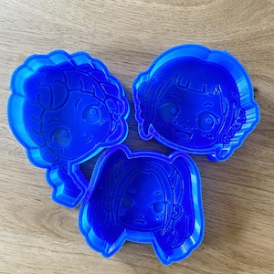May include: Three blue plastic cookie cutters in the shape of cartoon faces. The cutters have detailed facial features and different hairstyles. They are arranged on a light brown wooden surface.