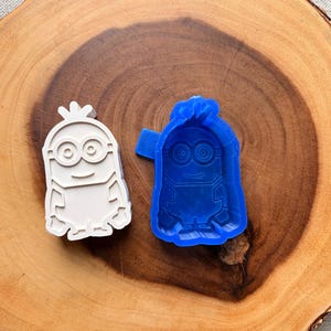 Peut inclure: Deux emporte-pièces en forme de Minion. L'un est blanc, l'autre bleu. Les emporte-pièces sont sur une surface en bois. Le design Minion présente de grands yeux, une bouche souriante et une petite touffe de cheveux.