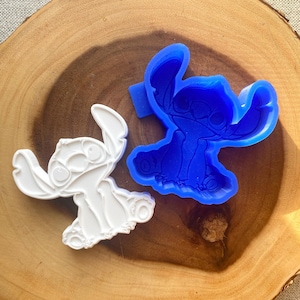 May include: Two Stitch-shaped molds. One is white, the other is blue. The molds are on a wooden surface. The white mold is a finished product, while the blue mold is a silicone mold. The molds are for crafting or baking.