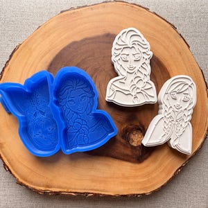May include: A wooden cutting board displays blue plastic molds and white cookie cutters. The molds are in the shape of two characters, and the cookie cutters are in the shape of two other characters. The characters have long braids and are from a popular animated movie.