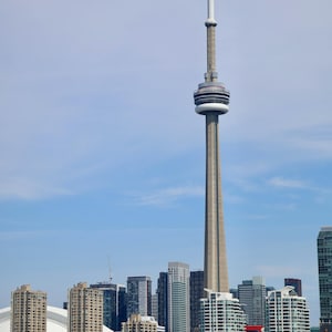 May include: A daytime shot of the Toronto skyline, featuring the CN Tower, a tall concrete structure with a distinctive observation deck. The tower stands prominently among various skyscrapers and buildings. Boats are visible on the water in the foreground.