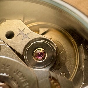 May include: Close-up of a watch mechanism with intricate details. The image showcases the internal components, including gears, jewels, and a balance wheel. The color palette is dominated by metallic tones, with a focus on precision engineering and craftsmanship.