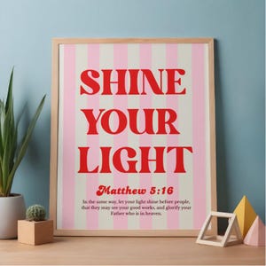 Könnte beinhalten: Ein gerahmter Druck mit den Worten "SHINE YOUR LIGHT" in Rot auf einem rosa-weiß gestreiften Hintergrund. Der Text "Matthew 5:16" und ein zugehöriges Zitat stehen darunter. Der Rahmen ist hellbraun.