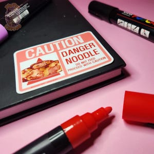 Może przedstawiać: Naklejka z napisem "CAUTION DANGER NOODLE" i ilustracją węża. Naklejka znajduje się na czarnym notatniku, z czerwonym markerem i innymi materiałami artystycznymi w tle. Na naklejce widnieje również napis "DO NOT FEED PROCEED WITH CAUTION."