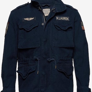 以下が含まれることがあります： ネイビーブルーのジャケット。複数のポケット、スタンドカラー、ウエストのドローコードが特徴です。「R.LAUREN」の文字とアメリカ国旗を含むパッチが付いています。ジャケットはジッパー開閉で、袖口はボタン留めです。