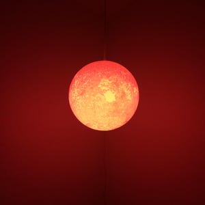 May include: A round, textured, red and yellow pendant light fixture. The light is suspended from a thin wire against a red background. The light's surface has a mottled appearance, with a bright yellow center.