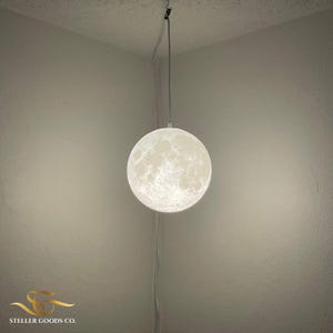 Puede incluir: Una lámpara esférica con forma de luna, blanca y texturizada, cuelga de un cable. La lámpara está contra una pared neutra. El accesorio de iluminación es un artículo de decoración del hogar único. El diámetro de la lámpara es de unos 30 cm.