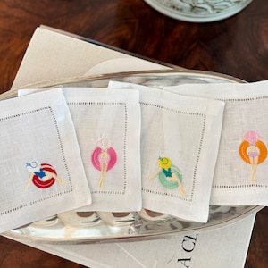 May include: Four square white linen cocktail napkins featuring embroidered figures floating in colorful inner tubes. Each napkin displays a different combination of a swimsuit and a pool float in red, pink, green, or orange, finished with a hemstitched border.