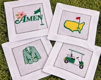 May include: Four white square coasters with embroidered golf-themed designs. One coaster features the word "Amen" with a flag and flower, another shows a map of the USA with a flag, a third has a green jacket, and the last depicts a golf cart.
