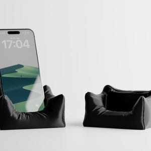 Könnte beinhalten: Zwei schwarze Telefonständer. Einer hält ein Smartphone mit einem Landschaftsbild und der Uhrzeit 17:04. Der andere Ständer ist leer. Beide Ständer haben ein weiches, kissenartiges Aussehen mit einem geschwungenen Design.