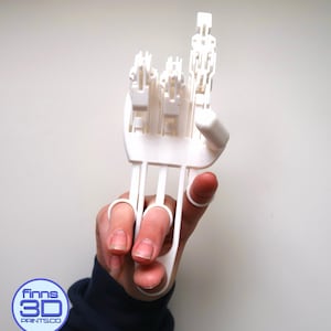 May include: A white 3D-printed hand model, with articulated fingers, is held up against a white background. The model is designed to fit over the fingers and thumb. The logo "finns3DPRINTS.co" is visible in the lower left corner.