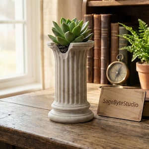 Puede incluir: Una pequeña maceta estriada de color blanquecino en forma de columna clásica, que contiene una suculenta verde. La maceta está sobre una superficie de madera, con un letrero de madera que dice "SageByteStudio". Libros y una brújula en el fondo.