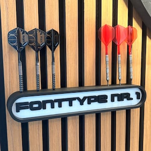Könnte beinhalten: Ein schwarz-weißer Wand-Darthalter mit der Aufschrift FONTTYPE NR. 1, der drei schwarze Darts mit gemusterten Flights auf der linken Seite und drei rote, durchscheinende Darts auf der rechten Seite in einer horizontalen Halterung präsentiert.