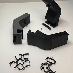 Könnte beinhalten: Schwarze Kamerabefestigungskomponenten und Clips sind auf einer weißen Oberfläche angeordnet. Die Komponenten umfassen mehrere gebogene Teile mit Kamerahalterungen, ein kleines rechteckiges Teil und eine Reihe kleiner schwarzer Clips.