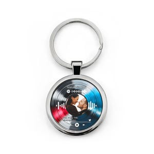 Op de afbeelding: Een zilveren sleutelhanger met een ronde hanger met een foto van een stel en een Spotify-code. De achtergrond heeft rode en blauwe accenten. De tekst "Sarah & John" en "Your Song Title" is zichtbaar.