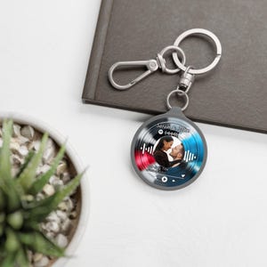 Puede incluir: Un llavero personalizado con la foto de una pareja y un código de Spotify. El colgante redondo tiene un borde gris con un diseño de gradiente rojo y azul. El llavero incluye un cierre y un anillo plateados.