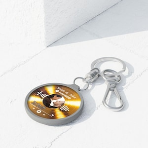 Puede incluir: Un llavero gris y dorado con un diseño circular. El llavero presenta una foto de una pareja besándose, los nombres "Sarah & John" y un código de Spotify. El llavero tiene un llavero y un cierre plateados.