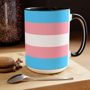 May include: A large ceramic mug featuring the transgender pride flag design. The mug has a white base, a black interior and handle, and a design of horizontal stripes in light blue, pink, and white. The mug is on a wooden surface with a spoon and a pastry.