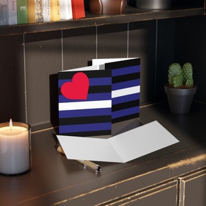 May include: Greeting cards with a red heart and a blue, black, and white striped design. One card is open, revealing a blank interior. A lit candle and a small potted cactus are in the background.