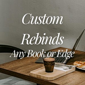 May include: A wooden table holds a laptop, a glass of dark coffee, and a small plate with food. The text "Custom Rebinds Any Book or Edge" is overlaid in white script font against the background, indicating a service for book restoration.