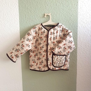 Könnte beinhalten: Cremefarbene Steppjacke mit einem Blumenmuster aus roten Blüten und grünen Blättern. Die Jacke hat eine dunkelbraune Paspel, eine Tasche und Holzknöpfe. Sie hängt an einem weißen Kleiderbügel vor einer zweifarbigen Wand.