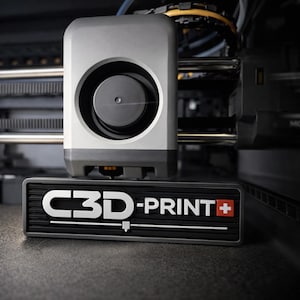 May include: A close-up of a 3D printer with a gray and black design. The printer features a prominent circular opening and the text "C3D-PRINT+" in white. The Swiss flag is incorporated into the design. The image is well-lit and showcases the printer's details.